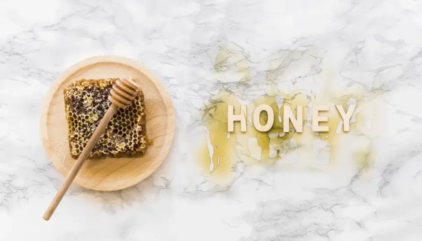 Plaster miodu z drewnianym mieszadełkiem na talerzyku i napis "HONEY" z miodu na marmurowym tle.